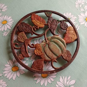 Peut inclure: Un dessous de plat rond en fonte représentant une citrouille et des feuilles. La citrouille est verte, et les feuilles sont de différentes nuances de marron, orange et jaune. Le dessous de plat a un aspect rustique et antique.
