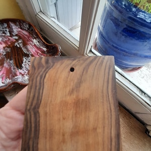 May include: A rectangular wooden cutting board with dark brown and light brown wood grain. The board has a small hole in the top center.