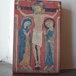 May include: A vintage-style religious artwork depicting a crucifixion scene. The central figure is Jesus Christ on the cross, with Mary and another woman standing on either side. The artwork is painted on a wooden panel with a red background and intricate details.