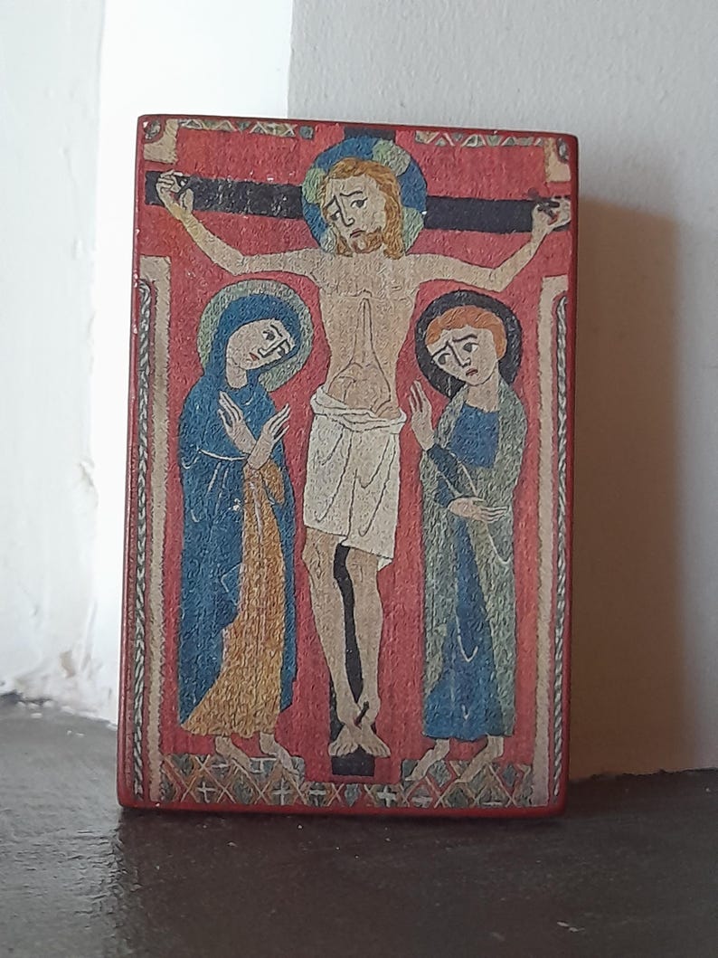 May include: A vintage-style religious artwork depicting a crucifixion scene. The central figure is Jesus Christ on the cross, with Mary and another woman standing on either side. The artwork is painted on a wooden panel with a red background and intricate details.