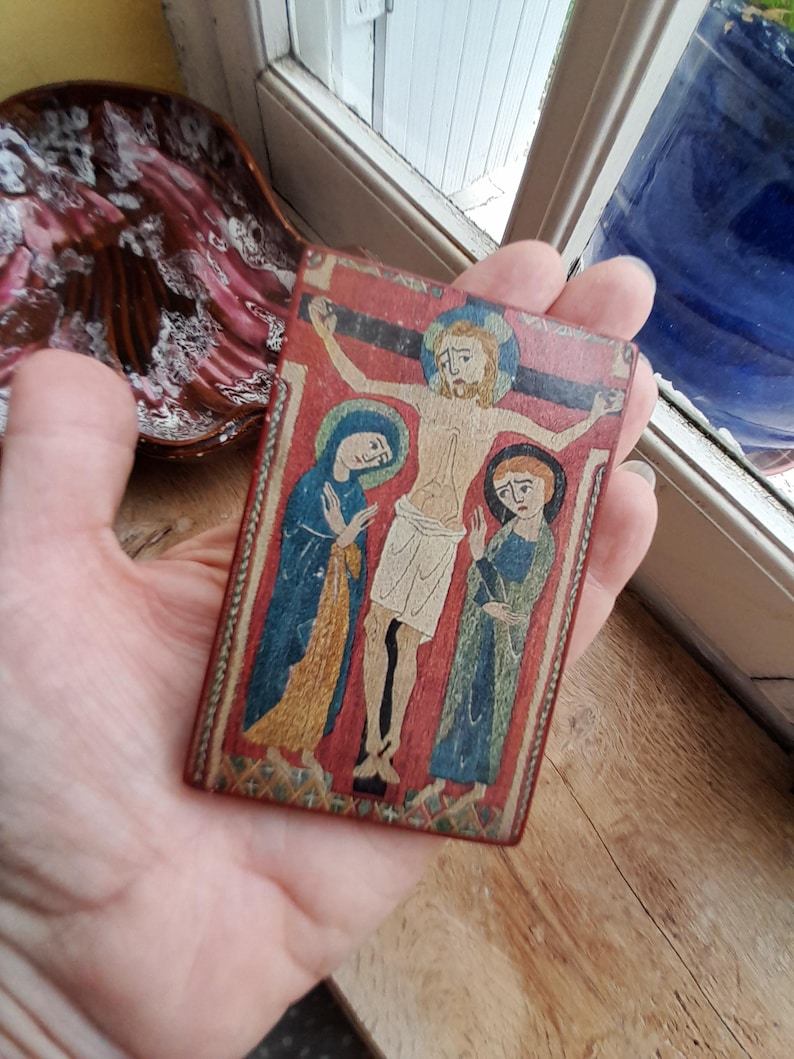May include: A small wooden plaque with a painted image of Jesus Christ on the cross. Two figures, likely Mary and Mary Magdalene, are flanking Jesus. The plaque has a red background with gold accents.