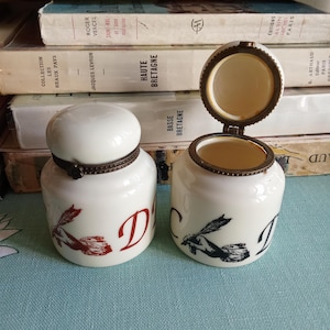 May include: Two white ceramic jars with lids, featuring a decorative design of a hand holding a feather and the letter "D". The jars have a brass-colored rim and are set against a backdrop of vintage books.