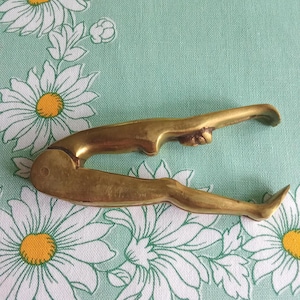 May include: A vintage brass nutcracker shaped like a reclining figure. The gold-toned nutcracker has a detailed design of a person with legs and arms. It rests on light green fabric with white daisy patterns.