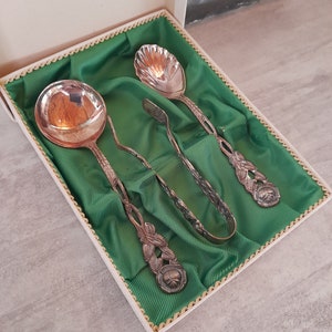 Pode incluir: Um conjunto de três pinças de açúcar e colheres prateadas em uma caixa forrada de veludo verde. As colheres têm desenhos florais ornamentados e as pinças têm um design simples.