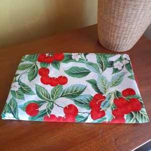 May include: A white fabric tablecloth with a red cherry print. The tablecloth features green leaves and white flowers. The tablecloth is folded and lying on a wooden table.