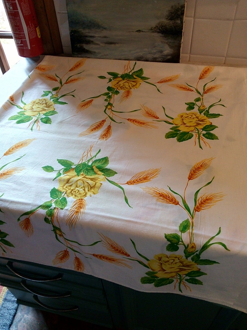 K&ouml;nnte beinhalten: Wei&szlig;e Tischdecke mit einem Muster aus gelben Rosen und Weizen. Die Tischdecke liegt auf einer dunkelgrauen Kommode mit zwei Schubladen.
