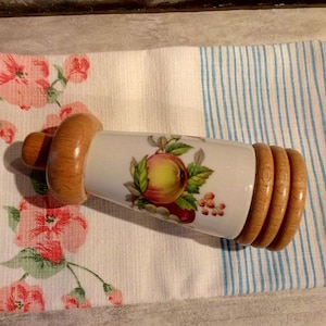 Peut inclure: Un bouchon de bouteille en céramique blanche avec un motif de fruits et un dessus en bois. Le bouchon de bouteille est posé sur un tissu rayé.