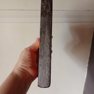 May include: A rectangular wooden block with a dark brown, weathered finish and a visible vertical grain texture. The object has rounded edges and a worn, rustic appearance, showing signs of age and natural wood characteristics throughout its surface.
