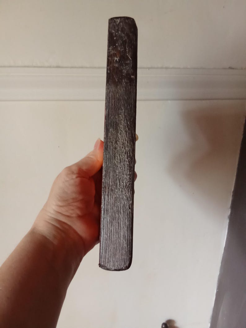 May include: A rectangular wooden block with a dark brown, weathered finish and a visible vertical grain texture. The object has rounded edges and a worn, rustic appearance, showing signs of age and natural wood characteristics throughout its surface.