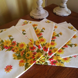 Könnte beinhalten: Satz von sechs weißen Baumwollservietten mit einem floralen Muster aus gelben und orangefarbenen Blumen auf einem braunen Holztisch.