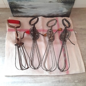 Puede incluir: Cuatro batidores de huevos giratorios vintage con varillas de metal y mecanismos de engranajes. El primer batidor presenta un mango y pomo de manivela estriados de color rojo brillante, mientras que los otros tienen mangos de metal o madera.