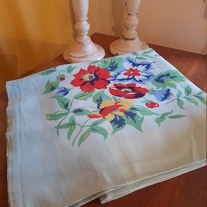 May include: A light blue tablecloth with a floral pattern featuring red, yellow, blue, and green flowers. The tablecloth is folded in half and sitting on a wooden table.