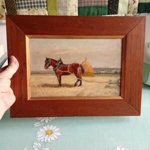 May include: A framed painting depicting a brown horse pulling a cart in a field. The horse is harnessed and stands near a haystack. The painting is in a wooden frame.