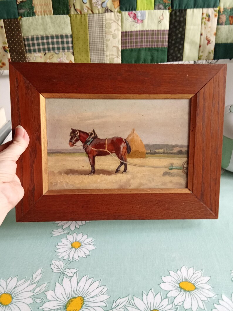 May include: A framed painting depicting a brown horse pulling a cart in a field. The horse is harnessed and stands near a haystack. The painting is in a wooden frame.