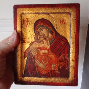 May include: A rectangular religious icon featuring a painted depiction of a figure holding a child against a gold leaf background. The border is painted in a deep red hue. Greek lettering is visible in the upper corners and on the left side.