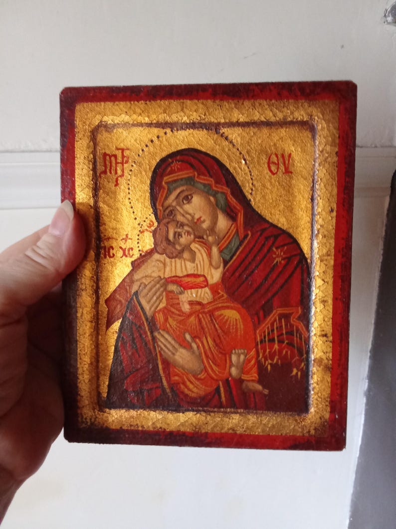 May include: A rectangular religious icon featuring a painted depiction of a figure holding a child against a gold leaf background. The border is painted in a deep red hue. Greek lettering is visible in the upper corners and on the left side.