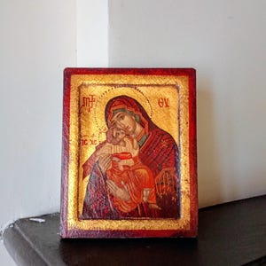 May include: A rectangular religious icon featuring a painted depiction of a figure holding a child against a gold leaf background. The border is painted in a deep red with a textured finish. Greek lettering is visible in the upper corners.