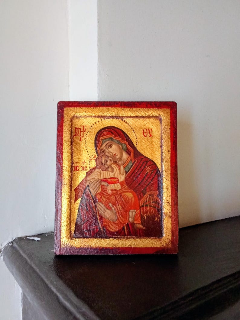 May include: A rectangular religious icon featuring a painted depiction of a figure holding a child against a gold leaf background. The border is painted in a deep red with a textured finish. Greek lettering is visible in the upper corners.