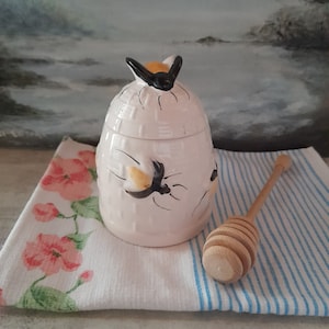 May include: A white ceramic honey pot shaped like a beehive with a bee on top. The pot has a honey dipper next to it on a striped blue and white cloth.