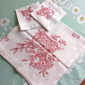 Pode incluir: Uma toalha de mesa de linho branco e duas bases para copos a condizer com desenhos de cestas de frutas bordadas a vermelho. A toalha de mesa e as bases para copos estão sobre uma superfície verde claro com padrão floral.