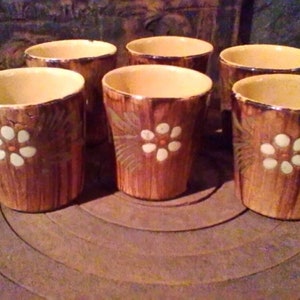 Puede incluir: Seis tazas de cerámica marrón con diseños de flores blancas. Las tazas tienen una textura rústica, similar a la madera.