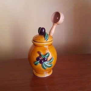 Op de afbeelding: Een keramische olijfolie dispenser met een houten lepel. De dispenser is versierd met een groene olijftak en zwarte olijven.