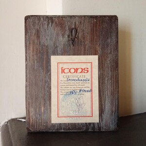 May include: A rectangular wooden plaque with a dark brown finish and visible wood grain. A metal hanging hook is attached to the top center. A rectangular certificate with a red border and the text "ICONS CERTIFICATE" is affixed to the center.