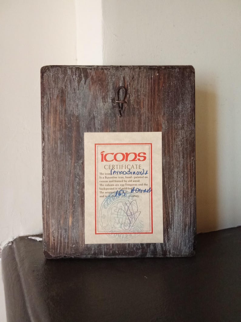 May include: A rectangular wooden plaque with a dark brown finish and visible wood grain. A metal hanging hook is attached to the top center. A rectangular certificate with a red border and the text "ICONS CERTIFICATE" is affixed to the center.