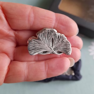 May include: A silver-toned metal pin shaped like a ginkgo leaf. The pin has a detailed design and is a delicate piece of jewellery.