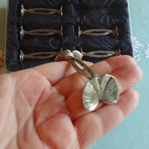 May include: A set of silver-toned napkin rings, one held in a hand, with a fan-like design. The rings are in a black velvet-lined box. The napkin ring in the hand has a leaf-like design.