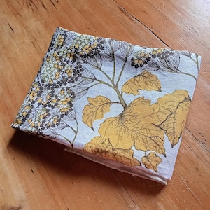 Puede incluir: Tela doblada con un estampado floral. El diseño presenta grandes hojas amarillas y grupos más pequeños de flores amarillas y marrones sobre un fondo azul claro. La tela está sobre una superficie de madera.
