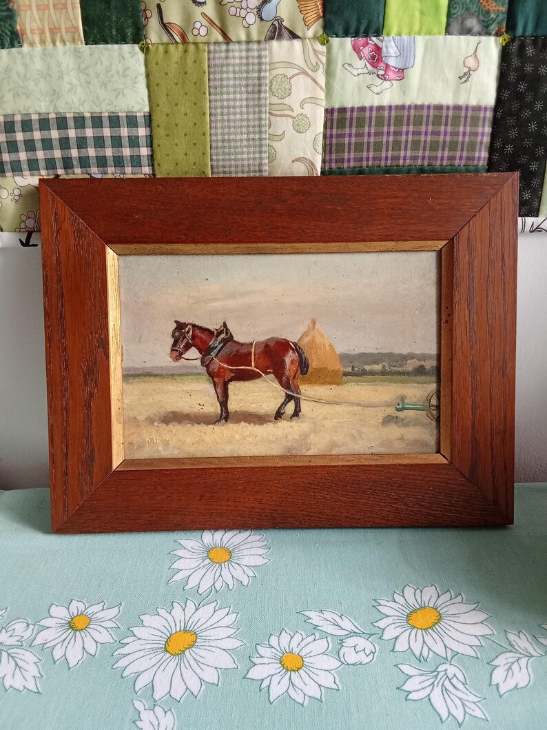 May include: A framed painting depicts a brown horse pulling a farm implement in a field. The horse is harnessed and connected to a piece of farm equipment. A haystack is in the background. The frame is made of dark wood.