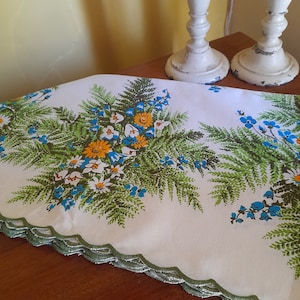 May include: White tablecloth with a scalloped edge and a floral print featuring blue, yellow, and green flowers. The tablecloth is on a wooden table.