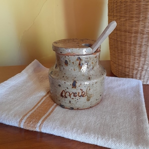 以下が含まれることがあります： 木製の蓋とスプーンが付いた、小さな斑点のある茶色の陶器の壺。壺には茶色の文字で「miel」とラベルが貼られています。