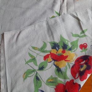 May include: A white tablecloth with a floral pattern featuring red, yellow, and blue flowers. The tablecloth has a green vine pattern and is made of a woven fabric.