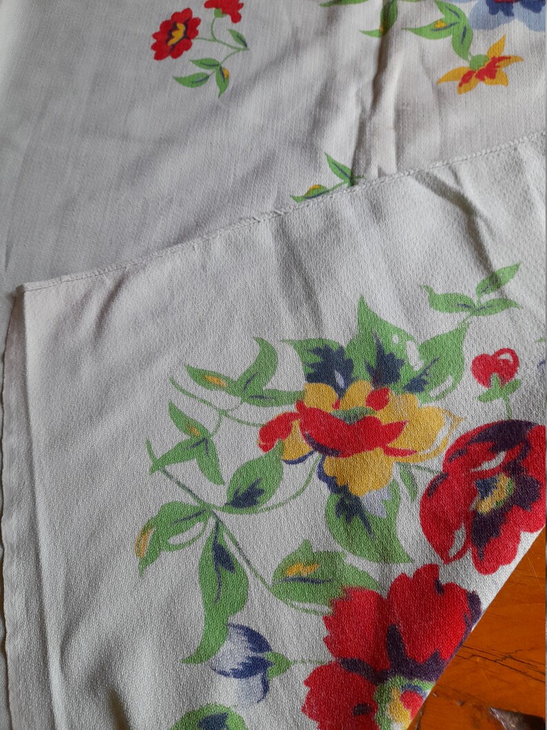 May include: A white tablecloth with a floral pattern featuring red, yellow, and blue flowers. The tablecloth has a green vine pattern and is made of a woven fabric.