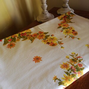 May include: White tablecloth with a floral pattern featuring yellow and orange flowers. The tablecloth is folded in half and lying on a wooden surface.