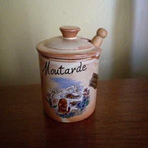May include: A ceramic mustard jar with a wooden lid and a painted scene of a mountain village. The jar is labeled "Moutarde" in black lettering.