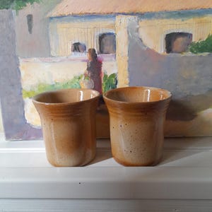 Könnte beinhalten: Zwei braune Keramikbecher mit einem gesprenkelten Überzug. Die Becher stehen auf einer weißen Oberfläche.