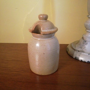 May include: A small, beige ceramic jar with a lid. The jar has a speckled glaze and a slightly rounded shape. The lid has a small opening for pouring.
