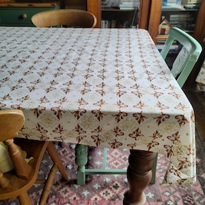 Puede incluir: Un mantel blanco con un patrón repetido de querubines dorados y estrellas. El mantel está puesto sobre una mesa de madera con dos sillas de madera. La mesa está en una habitación con un armario de madera en el fondo.