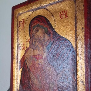 May include: A rectangular wooden icon featuring a painted image of a figure holding a child against a gold leaf background. The border is painted in a deep red color. Greek lettering is visible in the upper corners and near the child figure.