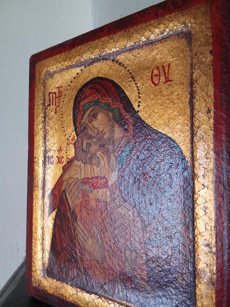 May include: A rectangular wooden icon featuring a painted image of a figure holding a child against a gold leaf background. The border is painted in a deep red color. Greek lettering is visible in the upper corners and near the child figure.