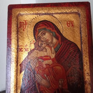 May include: A rectangular wooden icon featuring a painted depiction of a figure holding a child against a gold leaf background. The border is painted in a deep red hue. Greek lettering is visible in the upper corners and on the left side.