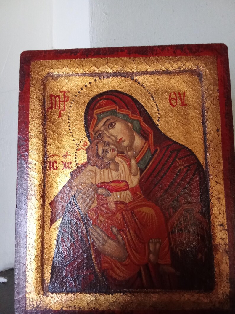 May include: A rectangular wooden icon featuring a painted depiction of a figure holding a child against a gold leaf background. The border is painted in a deep red hue. Greek lettering is visible in the upper corners and on the left side.
