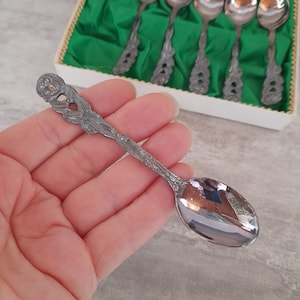 Pode incluir: Conjunto de seis colheres de chá de prata com desenhos florais ornamentados nas alças. As colheres estão em uma caixa branca com um forro de veludo verde.