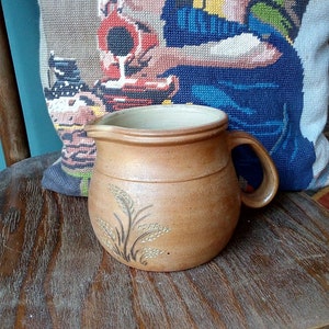 Puede incluir: Una jarra de cerámica marrón con un borde blanco y un diseño de espigas de trigo en el lateral. La jarra está sobre una superficie de madera.