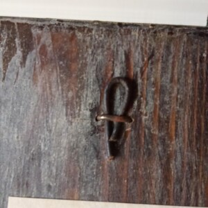 May include: A dark brown, weathered wooden surface featuring a vertical grain pattern. A small, dark, U-shaped metal wire loop is embedded into the center of the wood, serving as a fastener or hanging mechanism. The wood shows signs of age and wear.