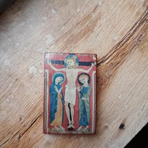 May include: A small wooden plaque with a painted image of Jesus Christ on the cross, flanked by two figures. The figures are painted in blue and yellow robes, with the background a red color.