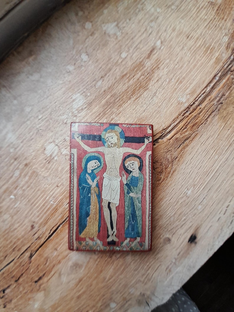 May include: A small wooden plaque with a painted image of Jesus Christ on the cross, flanked by two figures. The figures are painted in blue and yellow robes, with the background a red color.
